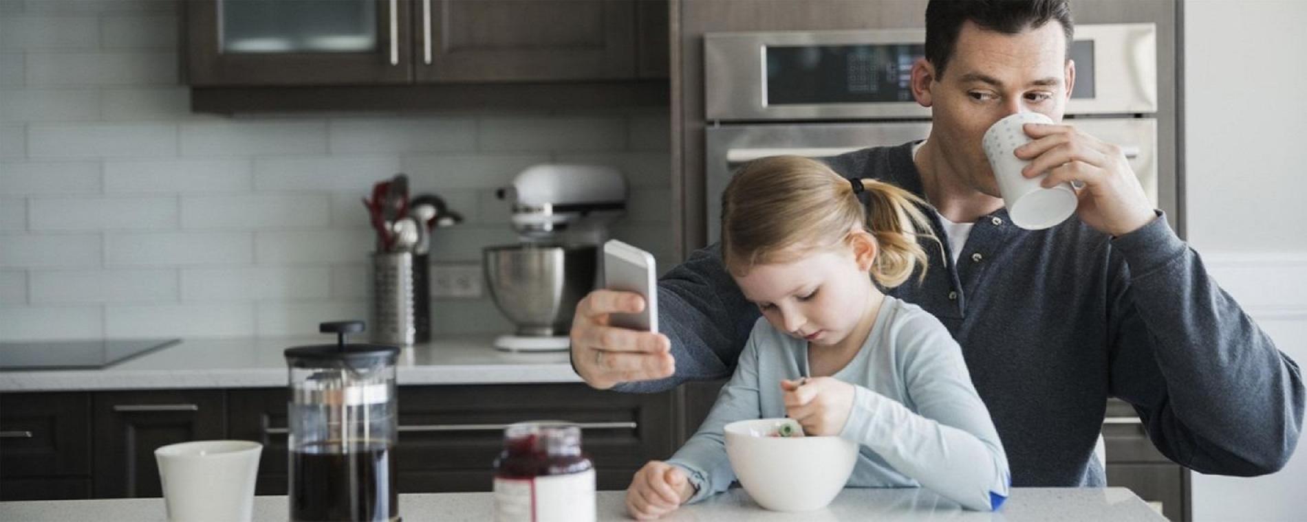 Busy Dad drinking coffee and checking his phone with his daughter on his lap.